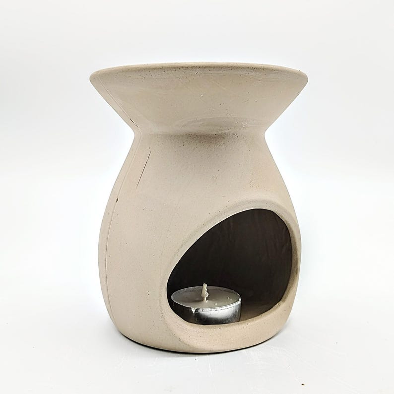May include: A beige ceramic oil burner with a round opening for a tea light candle. The burner has a wide, flat top and a rounded base.