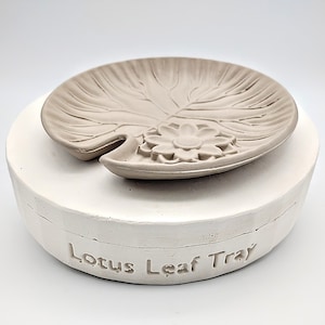 May include: A white ceramic mold for making a lotus leaf tray. The mold is in two pieces, with the top piece shaped like a lotus leaf with a flower in the center. The bottom piece is a round base with the words "Lotus Leaf Tray" inscribed on it.