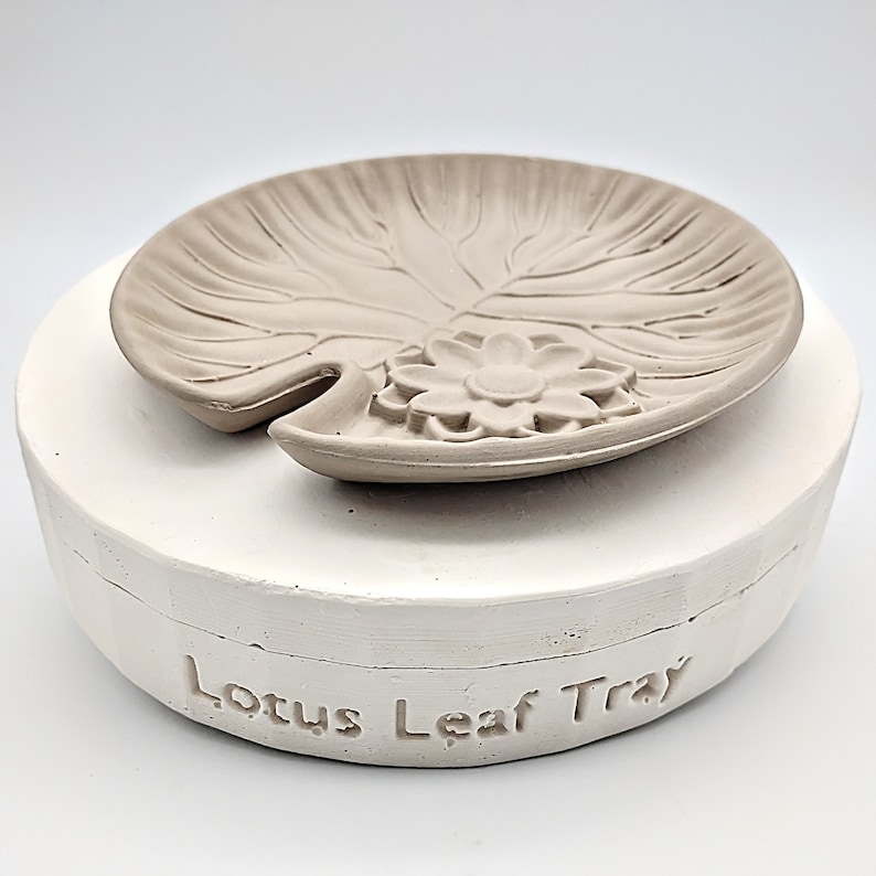 May include: A white ceramic mold for making a lotus leaf tray. The mold is in two pieces, with the top piece shaped like a lotus leaf with a flower in the center. The bottom piece is a round base with the words "Lotus Leaf Tray" inscribed on it.