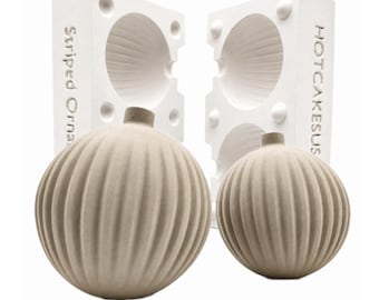 May include: Two beige, spherical, vertically ribbed ornaments positioned in front of two white rectangular molds. The left mold is embossed with the text Striped Orna, and the right mold features the text HOTCAKESUS along the side edge.