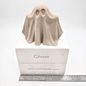 May include: A white ceramic mold for making a ghost-shaped cake. The mold is on a white base with the words "Ghost" and "HOTCAKESUSA.com" printed on it.
