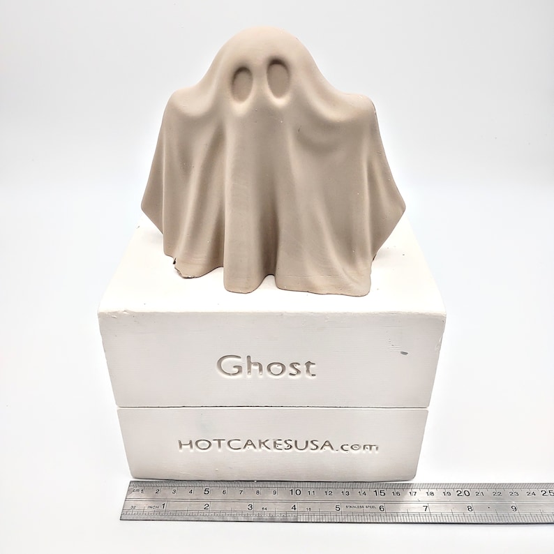 May include: A white ceramic mold for making a ghost-shaped cake. The mold is on a white base with the words "Ghost" and "HOTCAKESUSA.com" printed on it.