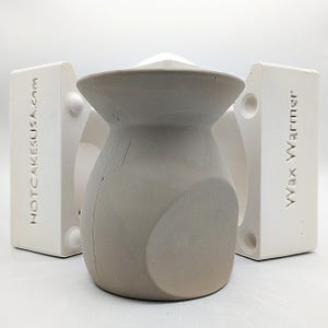 May include: A gray ceramic vase with a unique shape, sitting inside a white mold with the text "Wax Warmer" and "HOTCAKESUSA.com" on the sides.
