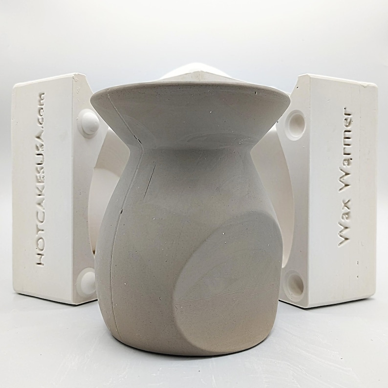 May include: A gray ceramic vase with a unique shape, sitting inside a white mold with the text "Wax Warmer" and "HOTCAKESUSA.com" on the sides.