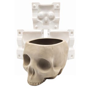 May include: A beige skull-shaped planter with a hollow interior and a smooth matte finish, positioned in front of three white rectangular casting molds that feature recessed skull impressions and circular alignment pegs on their edges.