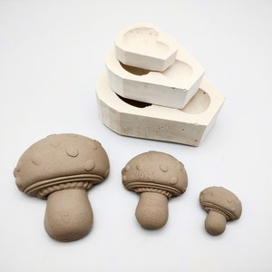 May include: Three brown ceramic mushroom figurines of varying sizes. The mushrooms have a textured surface with small dots and a raised rim around the cap. The mushrooms are arranged on a white surface.