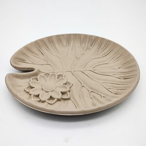 May include: A light brown ceramic dish shaped like a lily pad with a raised flower design in the center.