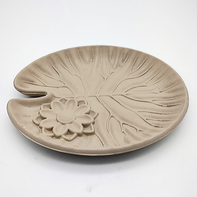May include: A light brown ceramic dish shaped like a lily pad with a raised flower design in the center.
