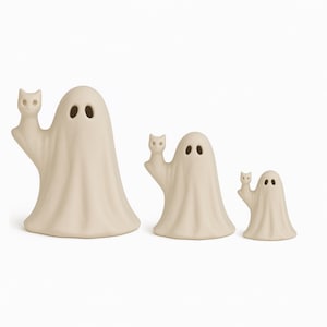 May include: Three off-white ceramic ghost figurines, each holding a small cat. The ghosts have oval eye cutouts and draped forms. The figurines vary in size, creating a decorative Halloween or fall theme.