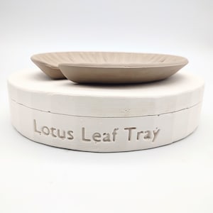 May include: A white ceramic tray with the words "Lotus Leaf Tray" embossed on the side. The tray has a matching beige ceramic lotus leaf dish on top.