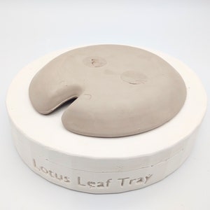May include: A light brown ceramic lotus leaf tray mold. The mold is sitting on a white base with the words "Lotus Leaf Tray" inscribed on the base.