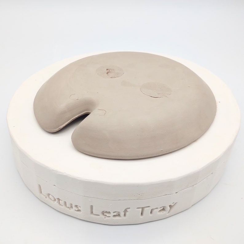 May include: A light brown ceramic lotus leaf tray mold. The mold is sitting on a white base with the words "Lotus Leaf Tray" inscribed on the base.