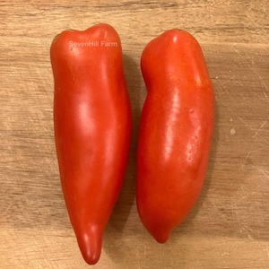 May include: Two bright red, elongated tomatoes on a wooden surface. The tomatoes are smooth and have a slightly pointed tip. The text "SevenHill Farm" is visible on the top of one tomato.
