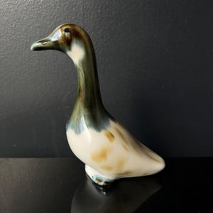 May include: A ceramic duck figurine with a long neck and a curved body. The duck has a white body with brown and tan markings. The neck is green and brown, and the beak is light blue. The figurine is on a black surface.
