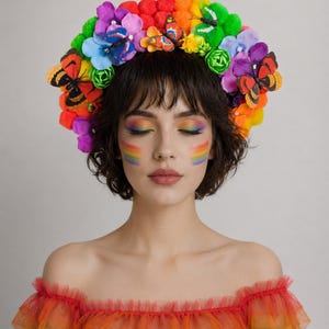 Könnte beinhalten: Ein Blumenkranz mit einer lebendigen Anordnung aus regenbogenfarbenen Pompons, künstlichen Blumen in Violett, Blau, Gelb und Orange sowie mehreren dekorativen Schmetterlingsornamenten, die über den gesamten kreisförmigen Kopfschmuck verteilt sind.