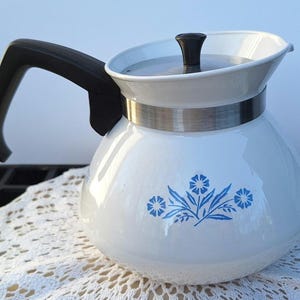 May include: A white teapot with a rounded body featuring a blue floral pattern. It has a black handle, a stainless steel band around the neck, and a matching white lid with a small black knob on top. The teapot has a wide spout and base.