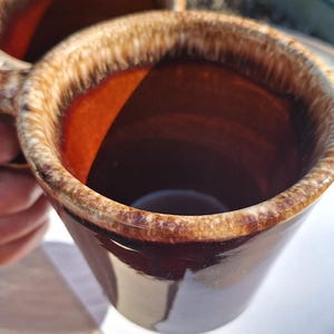 May include: A ceramic mug featuring a deep brown glossy glaze on the interior and exterior, complemented by a textured, lighter tan drip-style glaze around the rim. The mug has a sturdy handle and a cylindrical shape with a smooth, reflective finish.