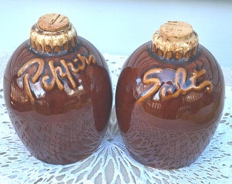 May include: Two brown glazed ceramic shakers with cork stoppers. One shaker features the word Pepper embossed in cursive script, and the other features the word Salt embossed in cursive script. Both have a textured, light-colored rim.