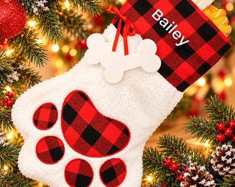 May include: A white plush paw-shaped Christmas stocking featuring a red and black buffalo plaid cuff with the name Bailey printed in white. The stocking includes a red ribbon tie with two white bone-shaped ornaments attached.
