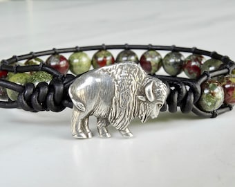 Può includere: Un braccialetto in pelle nera con perline di diaspro verde e rosso e un ciondolo a forma di bufalo d'argento.