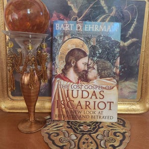 May include: A paperback book titled The Lost Gospel of Judas Iscariot by Bart D. Ehrman, featuring an illustration of two figures on the cover. Next to it stands a decorative metal candle holder with a large amber glass sphere on top.