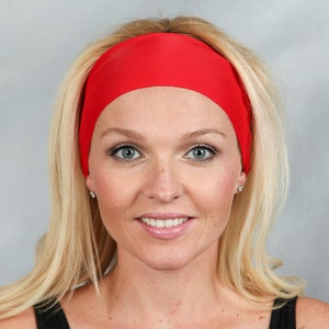 May include: A bright red headband worn across the forehead. The headband is made of a smooth, solid material. The person in the image has blonde hair and is wearing a black top.