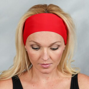 May include: A vibrant red headband, worn across the forehead. The headband is made of a smooth, solid material. The person in the image has blonde hair and is wearing a black top. The background is a solid light gray.