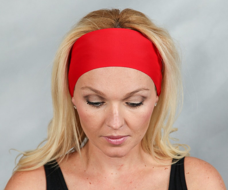 May include: A vibrant red headband, worn across the forehead. The headband is made of a smooth, solid material. The person in the image has blonde hair and is wearing a black top. The background is a solid light gray.