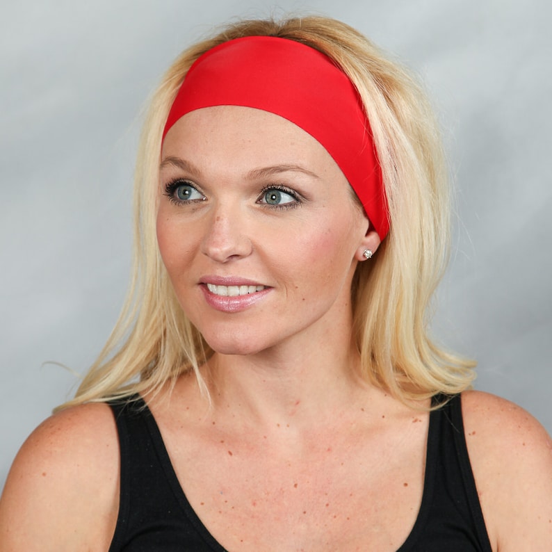 May include: A wide, solid red headband worn across the forehead. The headband is made of a smooth material and is worn by a person with blonde hair and a black tank top.