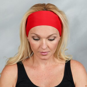 May include: A bright red headband worn by a person with blonde hair and a black tank top. The headband is wide and covers the forehead. The person is looking down with a neutral expression. The background is a solid light gray.