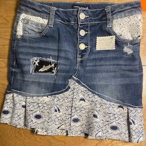 May include: Upcycled denim jean skirt with a shark-themed design. The skirt features a patchwork detail with a shark graphic, lace accents, and a pleated hem with a repeating shark pattern. The skirt has a button closure.