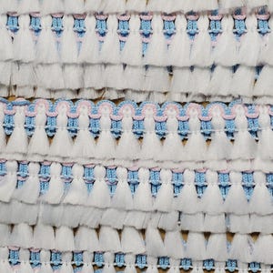 May include: A decorative trim featuring rows of white fringe tassels attached to a woven base. The base consists of light blue horizontal threads accented by a wavy, light pink braided cord pattern running horizontally across the fabric.