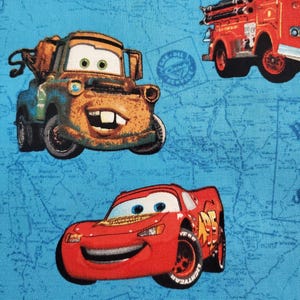 May include: A blue fabric with a map pattern featuring three cartoon cars. A red car with a number 95 on the side, a rusty brown tow truck, and a red fire truck.