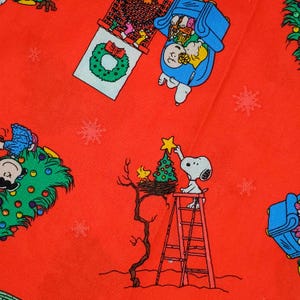 May include: Red fabric featuring a festive pattern of Snoopy and the Peanuts characters preparing for Christmas. Snoopy is on a ladder, placing a star on a Christmas tree. Other characters decorate a house and a Christmas tree. White snowflakes are scattered.