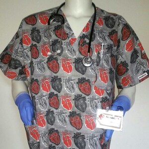 May include: Gray scrub top with a red and black anatomical heart print. The top has a V-neck and a pocket on the left side. A stethoscope is draped around the neck and a name tag with the text "ScrubL" is on the right side.
