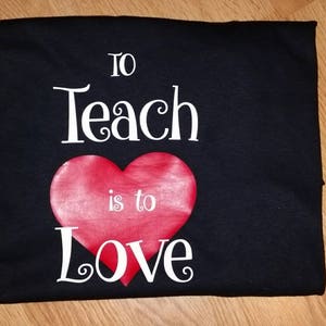 Pode incluir: Camiseta preta com a frase "To Teach is to Love" em escrita branca, com um cora&ccedil;&atilde;o vermelho no centro. A camiseta est&aacute; dobrada sobre uma superf&iacute;cie de madeira.