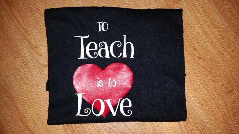 Pode incluir: Camiseta preta com a frase "To Teach is to Love" em escrita branca, com um cora&ccedil;&atilde;o vermelho no centro. A camiseta est&aacute; dobrada sobre uma superf&iacute;cie de madeira.