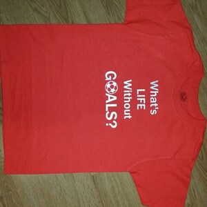 Op de afbeelding: Rood t-shirt met witte tekst "What's LIFE Without GOALS?" en een voetbalafbeelding. Het t-shirt is gemaakt van een zacht materiaal en is geschikt voor vrijetijdskleding. De tekst is vetgedrukt.