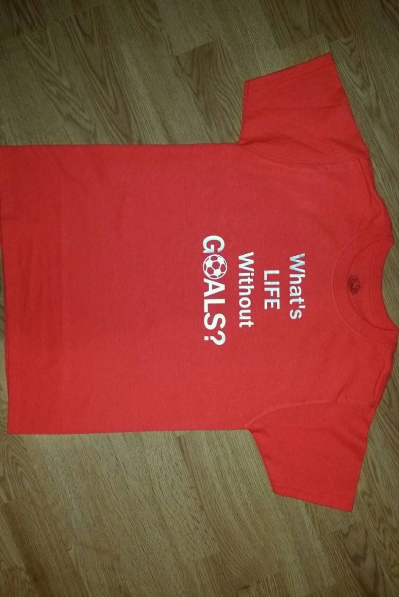Op de afbeelding: Rood t-shirt met witte tekst "What's LIFE Without GOALS?" en een voetbalafbeelding. Het t-shirt is gemaakt van een zacht materiaal en is geschikt voor vrijetijdskleding. De tekst is vetgedrukt.
