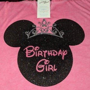 Może przedstawiać: R&oacute;żowa koszulka z czarną brokatową sylwetką Myszki Miki i srebrną brokatową koroną. Napis "Birthday Girl" jest w r&oacute;żowym skrypcie. Do koszulki przymocowana jest metka.