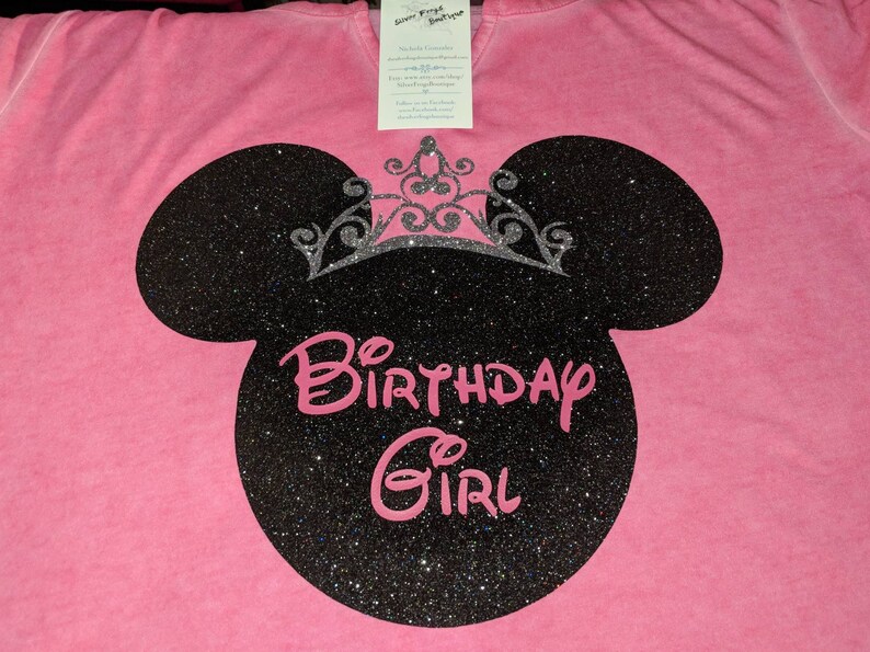 Może przedstawiać: R&oacute;żowa koszulka z czarną brokatową sylwetką Myszki Miki i srebrną brokatową koroną. Napis "Birthday Girl" jest w r&oacute;żowym skrypcie. Do koszulki przymocowana jest metka.