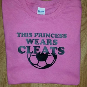 May include: A pink t-shirt featuring the text "THIS PRINCESS WEARS CLEATS" in black, with a black football graphic. The shirt is folded, likely cotton. The design suggests a sports theme.