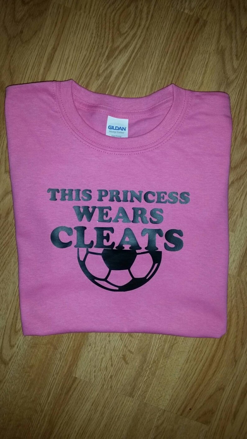 May include: A pink t-shirt featuring the text "THIS PRINCESS WEARS CLEATS" in black, with a black football graphic. The shirt is folded, likely cotton. The design suggests a sports theme.