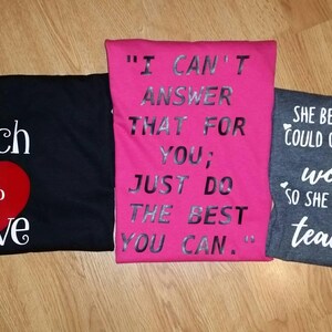 Pode incluir: Tr&ecirc;s camisetas dobradas com cita&ccedil;&otilde;es inspiradoras. Uma camiseta preta diz "To Teach is to Love" com um cora&ccedil;&atilde;o vermelho. Uma camiseta rosa diz "I Can't Answer That For You; Just Do The Best You Can." Uma camiseta cinza diz "She Believed She Could Change The World So She Became a Teacher."