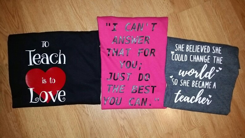 Pode incluir: Tr&ecirc;s camisetas dobradas com cita&ccedil;&otilde;es inspiradoras. Uma camiseta preta diz "To Teach is to Love" com um cora&ccedil;&atilde;o vermelho. Uma camiseta rosa diz "I Can't Answer That For You; Just Do The Best You Can." Uma camiseta cinza diz "She Believed She Could Change The World So She Became a Teacher."