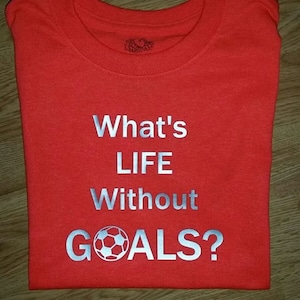 Op de afbeelding: Een opgevouwen rood t-shirt met de zilveren tekst "What's LIFE Without GOALS?" en een voetbalafbeelding. Het shirt is gemaakt van een zacht materiaal.