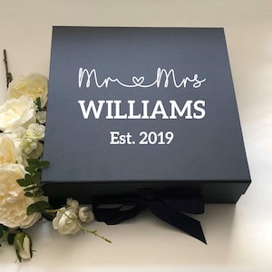 May include: A dark grey gift box with a white ribbon and a white inscription that reads "Mr & Mrs WILLIAMS Est. 2019".