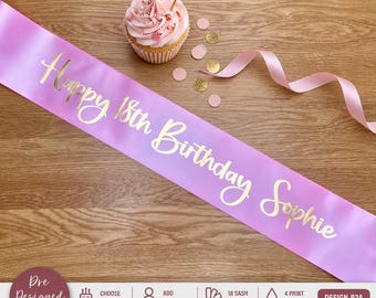 May include: A light purple satin sash featuring the gold foil text Happy 18th Birthday Sophie in a cursive script font. The sash is accompanied by a pink frosted cupcake topped with a small glittery heart pick and scattered confetti circles.