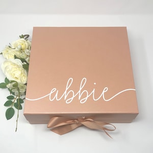 May include: A rose gold gift box with a white satin ribbon and the name "abbie" written in white cursive on the top.