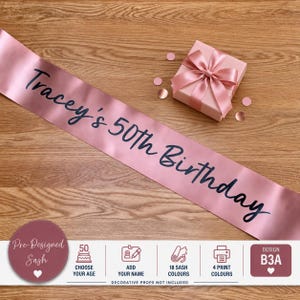 May include: A rose gold satin sash featuring the black text Tracey's 50th Birthday, accompanied by a small matching gift box tied with a satin ribbon and several small circular rose gold confetti pieces scattered nearby.